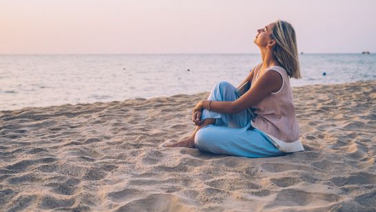 Junge Frau sitzt entspannt am Strand und schaut aufs Meer - Foto: canva.com