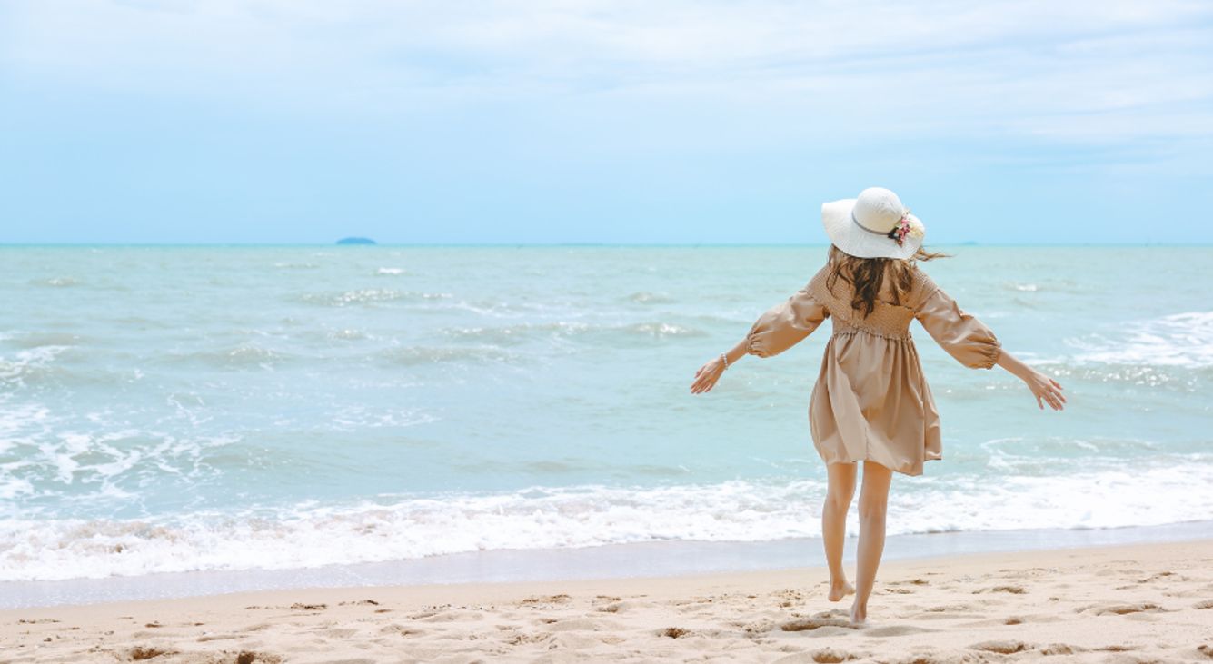 Frau im Sommerkleid steht am Strand und schaut auf das blauen Meer