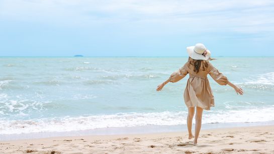 Frau im Sommerkleid steht am Strand und schaut auf das blauen Meer - Foto: canva