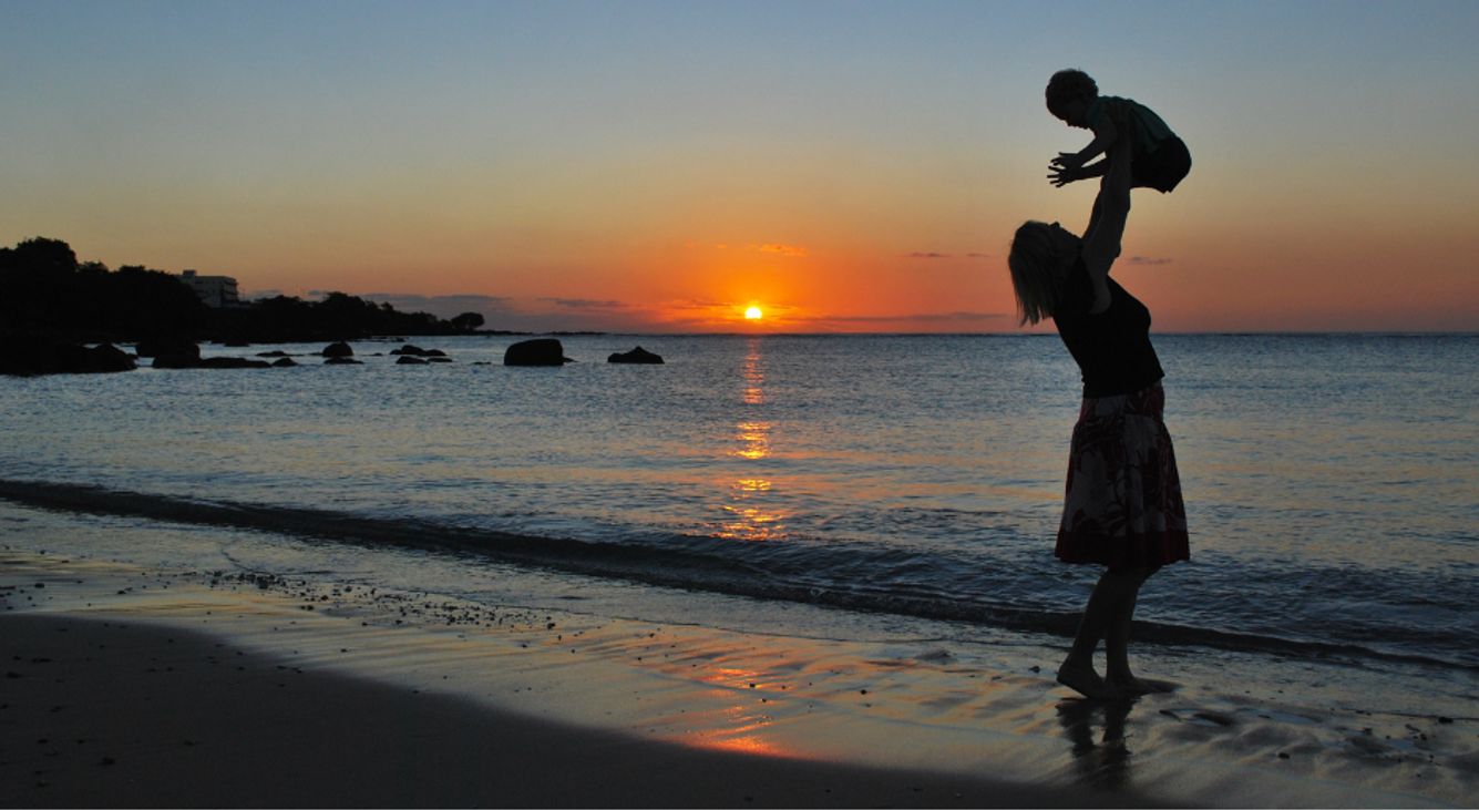 Mutter wirft ihr am Strand mit untergehender Sonne in die Luft und fängt es in ihren Armen wieder auf.