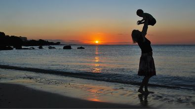 Mutter wirft ihr am Strand mit untergehender Sonne in die Luft und fängt es in ihren Armen wieder auf. - Foto: canva.com