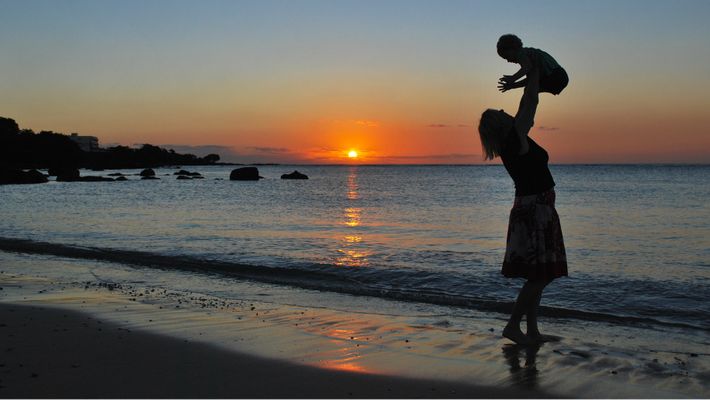 Mutter wirft ihr am Strand mit untergehender Sonne in die Luft und fängt es in ihren Armen wieder auf. - Foto: canva.com