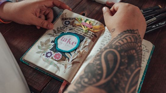 Eine Frau beugt sich über ein offenes Notizbuch mit Zeichnungen darin - Foto: canva.com