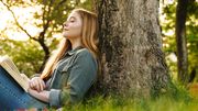 Frau mit Buch lehnt an Baum - Foto: canva.com