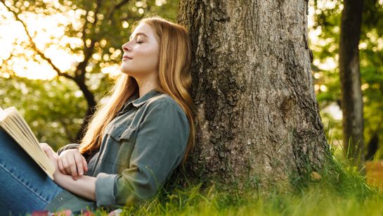 Frau mit Buch lehnt an Baum - Foto: canva.com